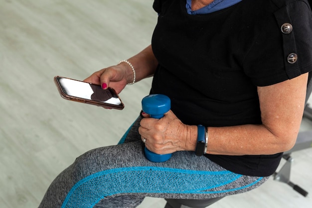 Una donna latina più anziana si allena in palestra con i manubri usando i video di allenamento del cellulare