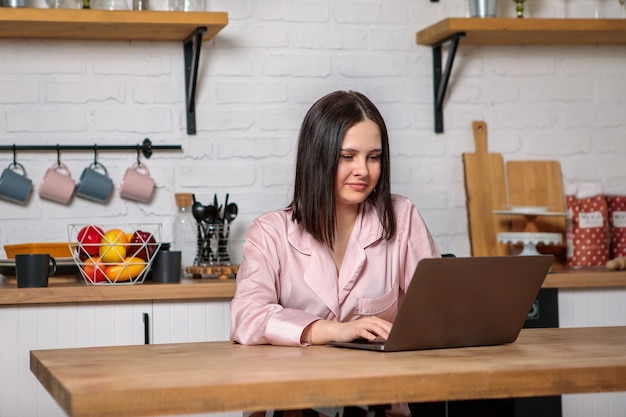 Una donna lavora in ufficio a distanza da casa in cucina, seduta al computer. Formazione e lavoro online a distanza. Studentessa si prepara per gli esami.