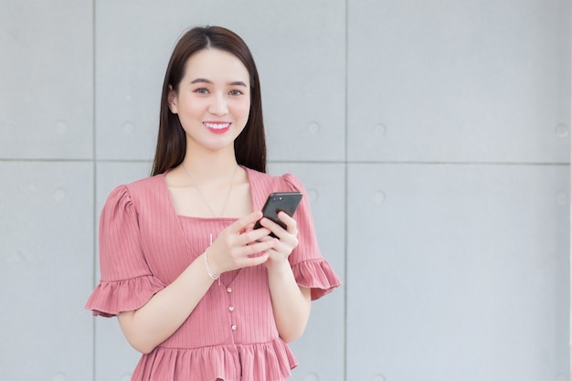 Una donna lavoratrice asiatica che ha i capelli lunghi e indossa un abito rosa tiene in mano uno smartphone e sorride