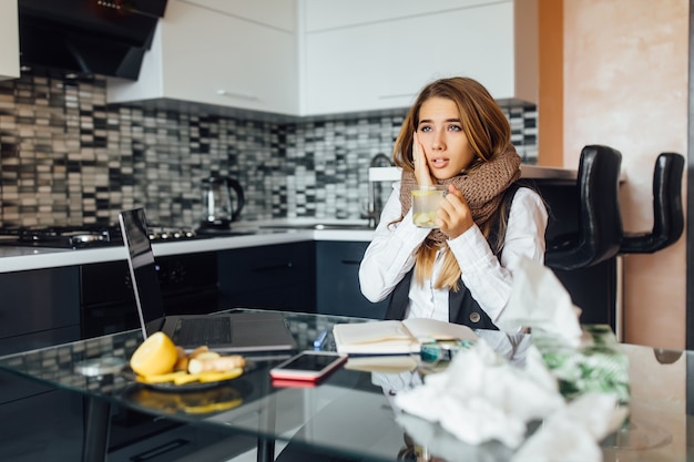 Una donna malata con una sciarpa calda seduta al tavolo in cucina tiene una tazza di tè al limone