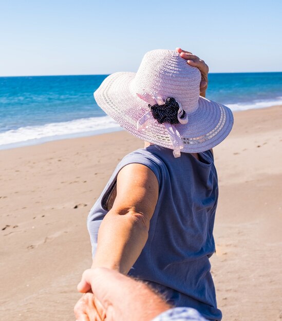 Una donna matura che cammina nella sabbia della spiaggia con suo marito che le tiene la mano - vacanze e vacanze all'aperto con pensionati di turisti che si divertono e si divertono