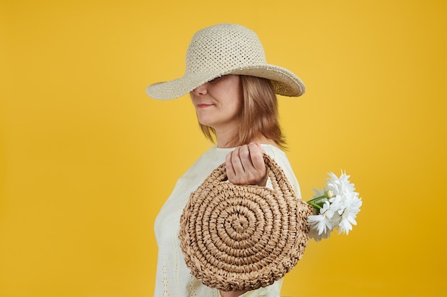 Una donna matura con una borsa di paglia si è tirata il cappello sul viso su uno sfondo giallo brillante.