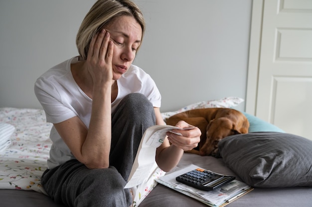 Una donna matura sconvolta con le bollette paga e la calcolatrice si siede sul letto con il cane nell'interno di casa m