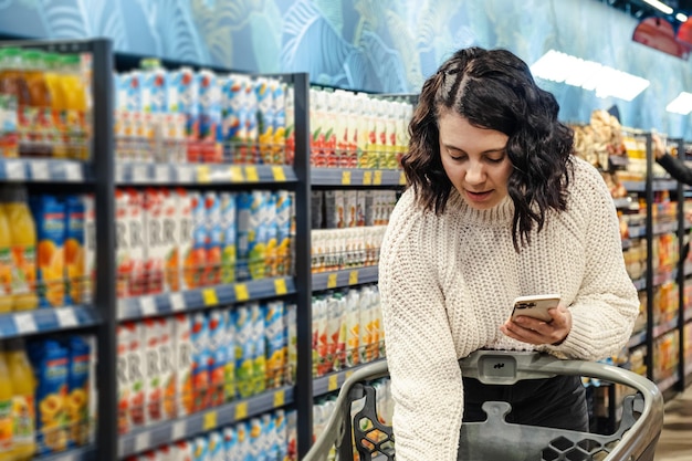 Una donna mette un prodotto con un elenco di prodotti sul telefono in un carrello del supermercato