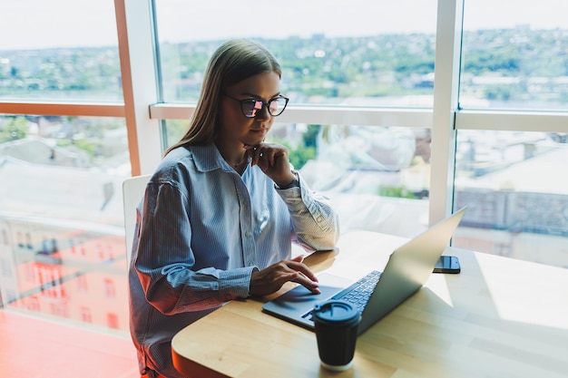 Una donna moderna guarda video su un laptop e beve caffè Il concetto di distanza o elearning Una giovane donna sorridente si siede su una sedia al tavolo