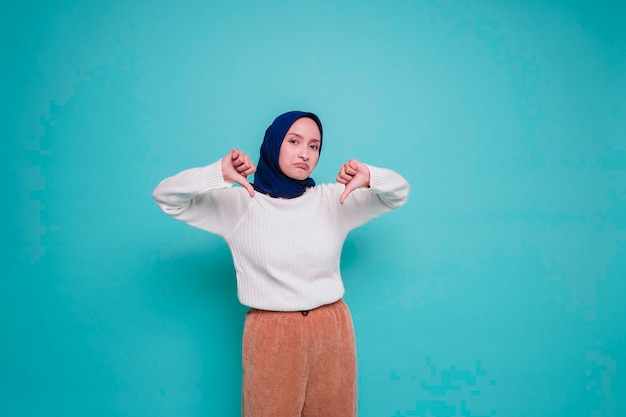Una donna musulmana asiatica delusa che indossa camicia bianca e hijab dà il pollice in giù gesto di approvazione della mano isolato da sfondo blu