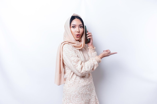 Una donna musulmana asiatica scioccata che indossa l'hijab indicando lo spazio della copia accanto a lei mentre tiene il telefono isolato da uno sfondo bianco