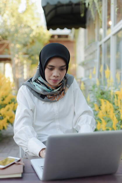 Una donna musulmana asiatica siede in un giardino con un computer portatile e un telefono in grembo