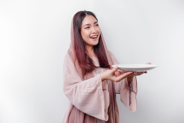 Una donna musulmana asiatica sorridente è a digiuno e affamata e tiene in mano e indica un piatto