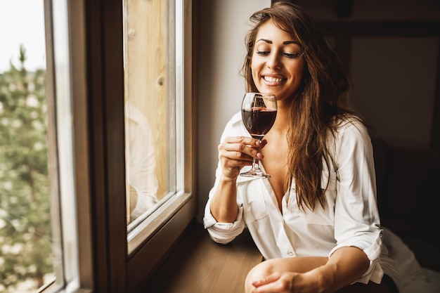 Una donna pensierosa e sorridente carina sta bevendo vino rosso mentre è in piedi vicino alla finestra e si diverte a casa.