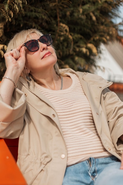 Una donna piuttosto anziana in abiti alla moda indossa occhiali da sole si siede su una terrazza e gode di una buia giornata primaverile