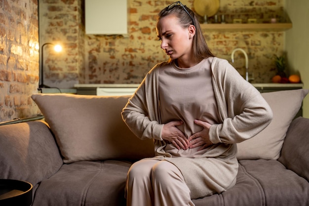 Una donna preoccupata che soffre di dolori addominali è seduta sul divano Una donna che si tiene la pancia provando dolore e fastidio all'addome soffre di mestruazioni