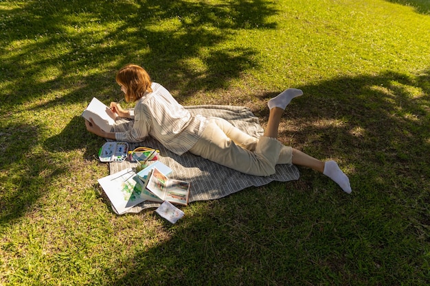 Una donna rilassata adulta sdraiata sulla coperta nel parco e disegnando nel suo taccuino