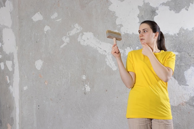 Una donna riparatrice con una maglietta gialla brillante tiene un pennello e guarda un muro vuoto con posto per il testo Riparazioni chiavi in mano