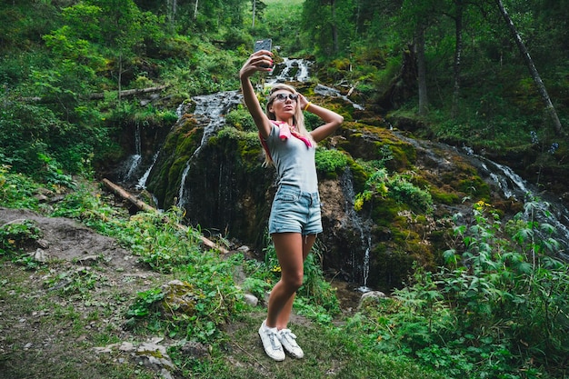 Una donna scatta una foto in natura sullo sfondo di una cascata Bella ragazza dai capelli lunghi nel fare selfie sullo sfondo di una cascata di fiume di montagna Sera d'estate