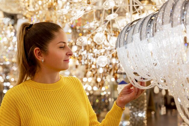 Una donna sceglie un bellissimo lampadario costoso nel salone Giovane bionda in un maglione giallo