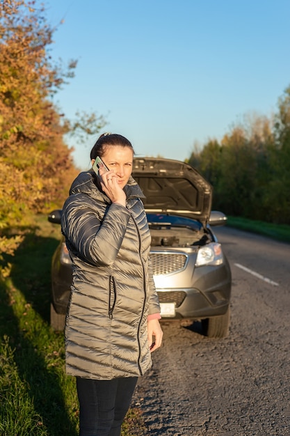 Una donna sconvolta sta vicino alla sua macchina rotta e parla al cellulare per strada in autunno.