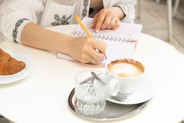 Una donna scrive con una penna in un quaderno seduta in un caffè una tazza di caffè sul tavolo mani in primo piano