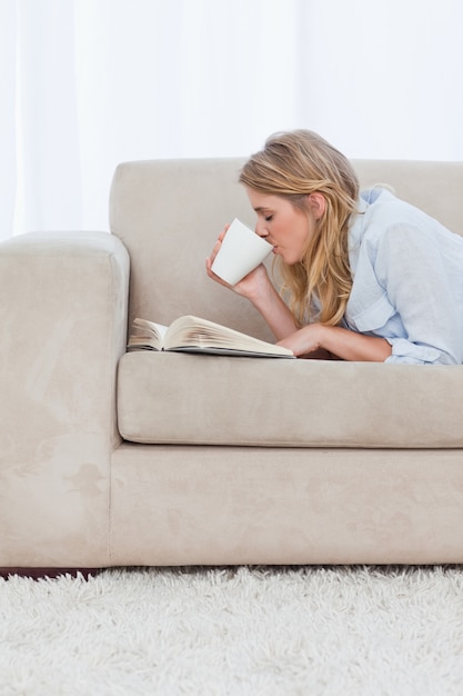 Una donna sdraiata su un divano con un libro sta bevendo un caffè