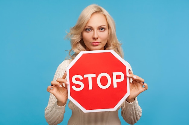 Una donna seria e concentrata con i capelli biondi che tiene in mano il segnale di stop, avvertendo del pericolo, nega le cattive abitudini. Studio indoor girato isolato su sfondo blu