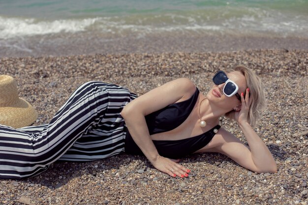 Una donna sexy in costume da bagno nero, pantaloni a righe e occhiali da sole si trova sulla spiaggia in riva al mare in estate