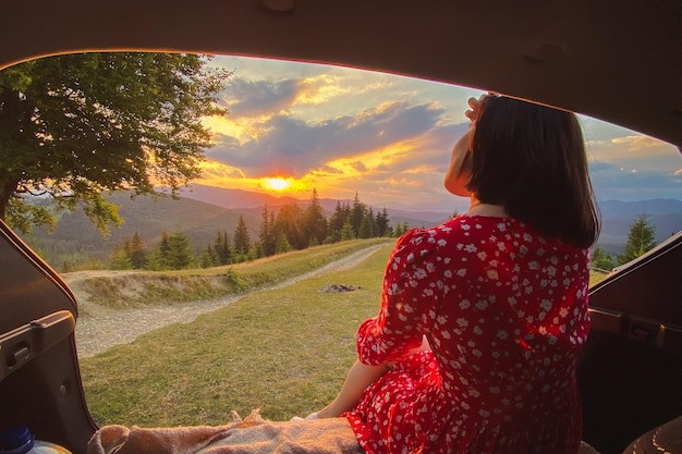 Una donna siede nel bagagliaio di un'auto con vista sul tramonto in montagna