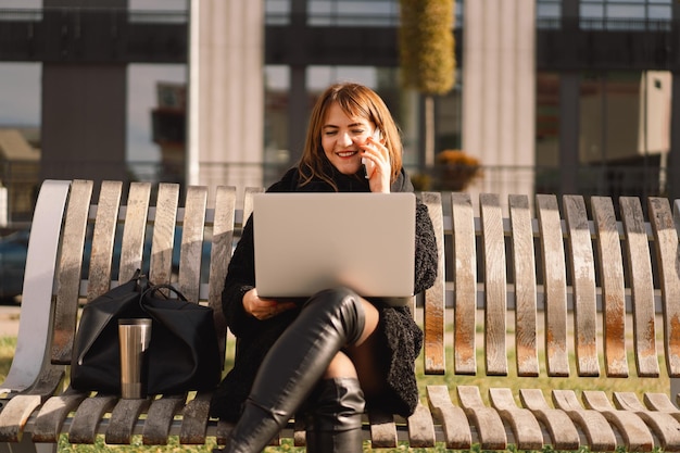 Una donna siede su una panchina in città, usa un computer e un telefono. Donna libera professionista. Persone e tecnologia