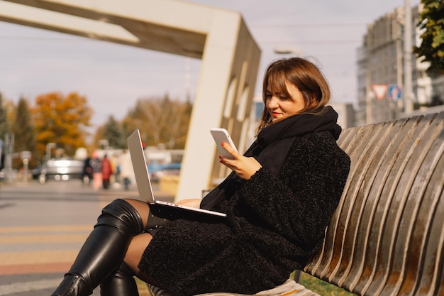 Una donna siede su una panchina in città, usa un computer e un telefono. Donna libera professionista. Persone e tecnologia