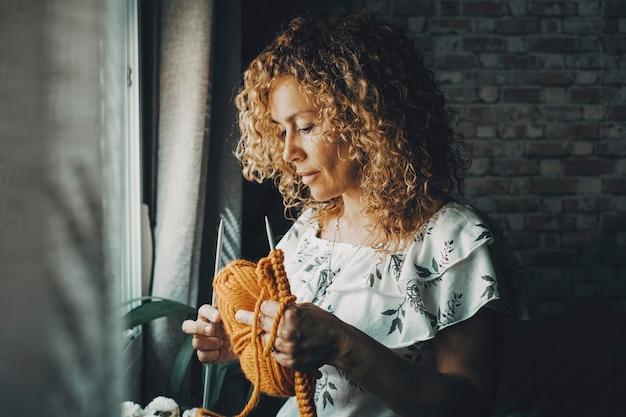 Una donna sola a casa impegnata a lavorare a maglia per hobby vicino alla finestra Attività per il tempo libero al chiuso persone di sesso femminile Appartamento luce scura sullo sfondo Femmina adulta che pensa e lavora per piacere