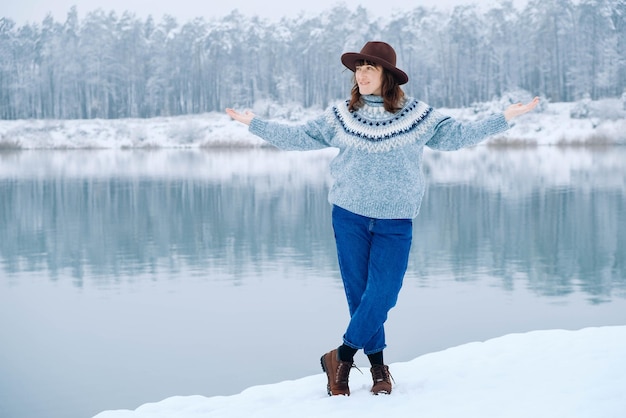 Una donna sorridente con un cappello marrone e un maglione si trova sulle rive di un lago e di una foresta innevati