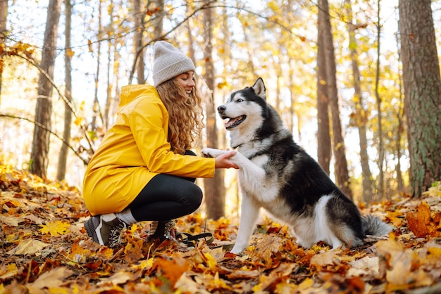 Una donna sorridente con un cappotto giallo cammina con il suo simpatico animale domestico Husky nella foresta autunnale con tempo soleggiato