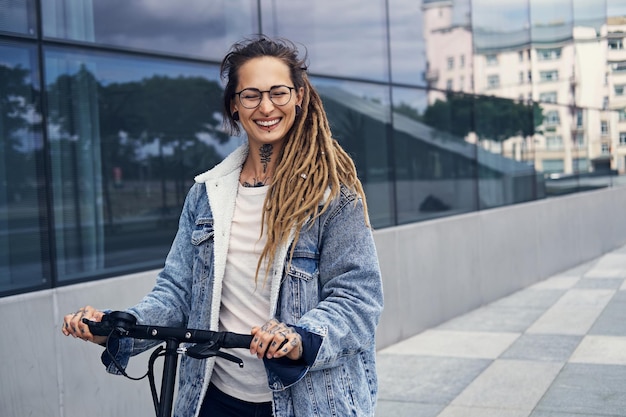 Una donna sorridente felice con tatuaggi e dreadlocks sta guidando uno scooter elettrico per strada.