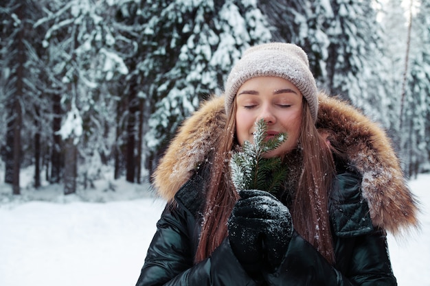 Una donna sorridente felice in abiti caldi tiene in mano un ramoscello di pino e inala l'aroma degli aghi di pino. Bella pineta sotto la neve, gelido inverno.