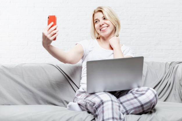 Una donna sorridente in pigiama si siede su un divano con un computer portatile e scatta una foto su uno smartphone. Blog e telelavoro.