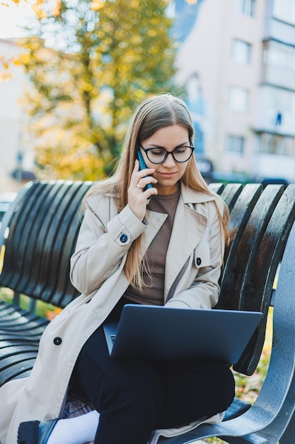 Una donna sorridente lavora con un laptop su una panchina nel Parco d'Autunno Ha un bel sorriso capelli lunghi e grandi occhi azzurri Ritratto di una donna che lavora moderna Sfondo giallo del parco