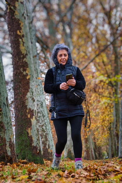 Una donna sorridente matura che cammina attraverso una foresta autunnale usando il suo telefono cellulare.