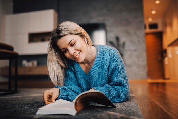 Una donna sorridente sdraiata sul pavimento nella sua casa accogliente e leggendo un libro