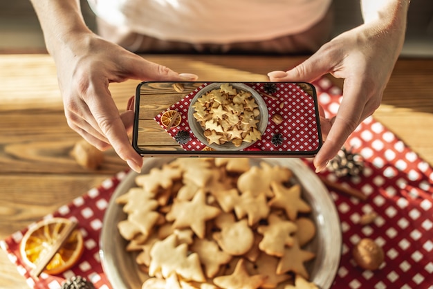 Una donna sta scattando foto un piatto con deliziosi biscotti natalizi fatti in casa sulla fotocamera del suo telefono