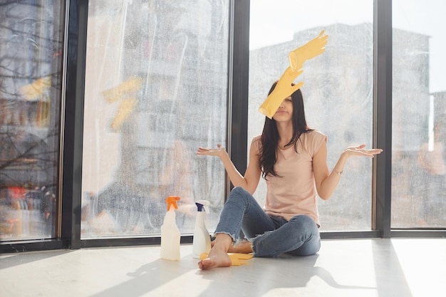 Una donna stanca si siede sul davanzale della finestra con bottiglie di spray detergente e lancia guanti in aria