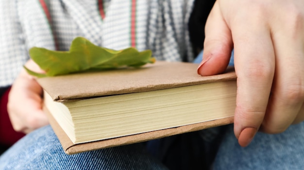 Una donna tiene un libro chiuso sdraiato in grembo con un primo piano di una foglia di quercia caduta in un parco in una calda giornata di sole autunnale. Il concetto di relax, lettura e relax da solo.