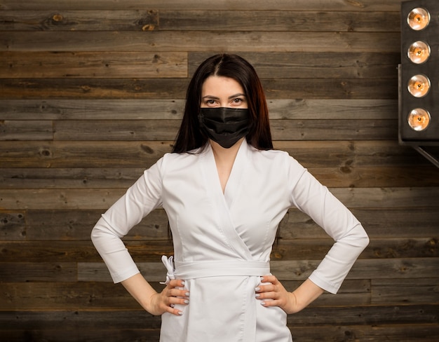 Una dottoressa in uniforme bianca e maschera medica nera si trova sulla parete di legno