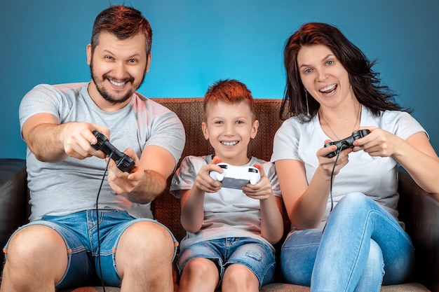 Una famiglia allegra, papà mamma e figlio giocano sulla console, i videogiochi, reagiscono emotivamente seduti sul divano. Giorno libero, divertimento, svago, trascorrere del tempo insieme.