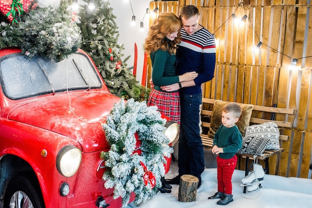 Una famiglia felice si diverte, gioca con il figlio piccolo vicino a un'auto rossa con regali, alberi di Natale