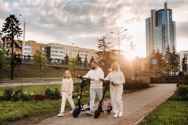 Una famiglia in abiti bianchi cammina per la città con scooter elettrici al tramonto. Attività all'aperto.