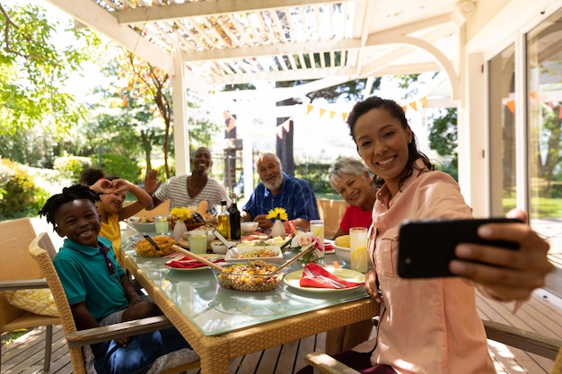 Una famiglia multietnica e multigenerazionale seduta a tavola per un pasto insieme all'aperto in un patio al sole, la madre di razza mista usa uno smartphone per scattare un selfie con tutti loro sorridenti