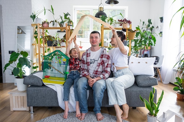 una famiglia tradizionale con un bambino e un pappagallo in gabbia si gode una nuova casa seduta sul divano e tiene il tetto Acquistando un'assicurazione ipotecaria e traslocando nella propria casa