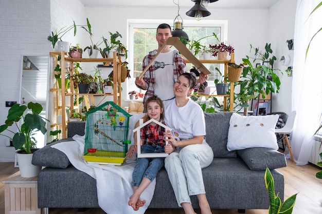 una famiglia tradizionale con un bambino e un pappagallo in gabbia si gode una nuova casa seduta sul divano e tiene il tetto Acquistando un'assicurazione ipotecaria e traslocando nella propria casa