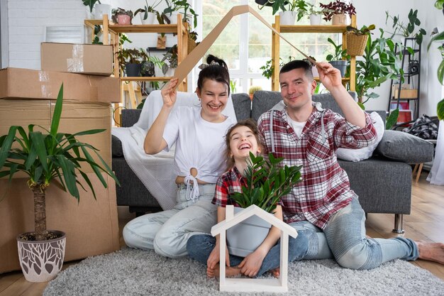 Una famiglia trasferita con un bambino e una pianta domestica si sta godendo una nuova casa seduta sul divano e tenendo il tetto vicino a scatole con cose Acquistando un'assicurazione ipotecaria e traslocando nella tua abitazione