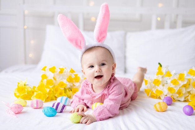 Una felice bambina caucasica con un bordo di orecchie da coniglio sulla testa giace sul letto di casa con uova di Pasqua colorate e fiori gialli primaverili guarda la telecamera ride il bambino di Pasqua