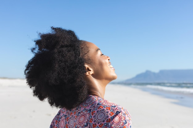 Una felice donna afroamericana che sorride sulla spiaggia soleggiata vicino al mare. Estate, benessere, libertà, relax e vacanze, inalterate.